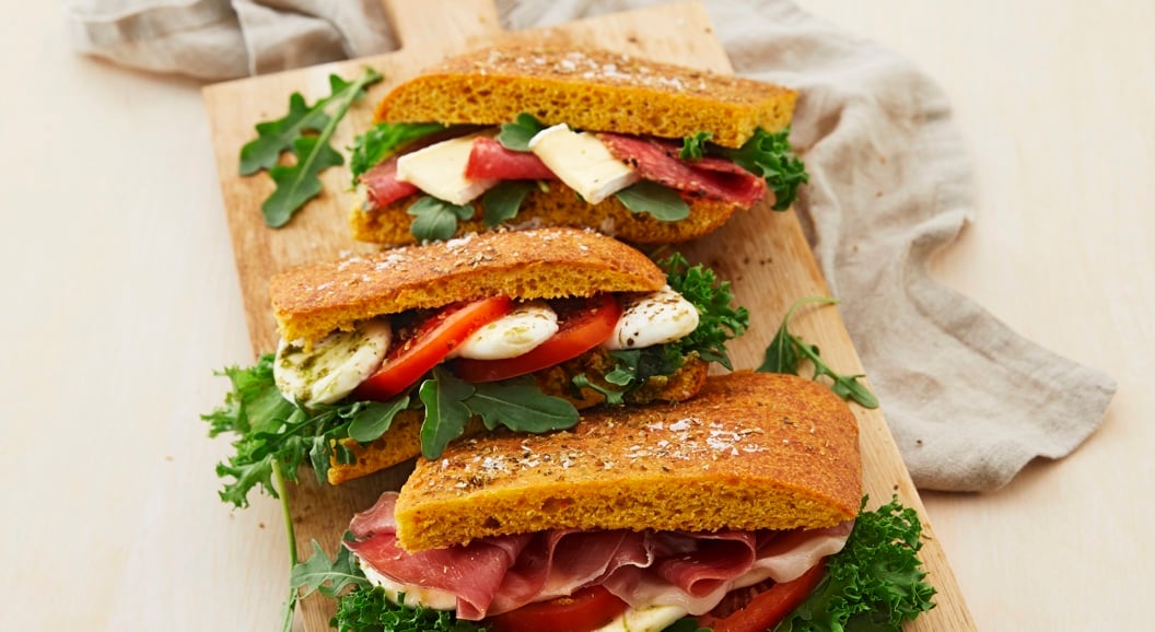 Three gourmet sandwiches with prosciutto, cheese, tomato, and arugula on a wooden board, styled with a linen cloth