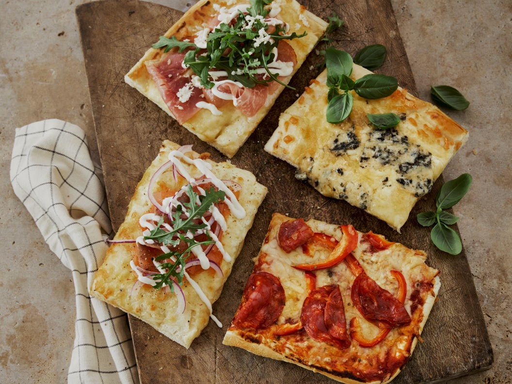 Fire stykker italiensk pizza på et rustikt trefat, hver med forskjellig topping som spekeskinke med ruccola, blåmuggost, løk med rømme, og pepperoni med paprika