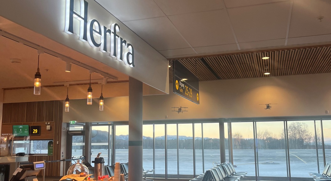 Modern airport waiting area with a cafe sign reading "Herfra" and large windows overlooking a runway