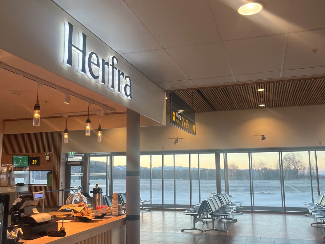 Modern airport waiting area with a cafe sign reading "Herfra" and large windows overlooking a runway
