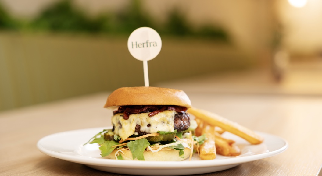 Gourmet cheeseburger on a white plate with fries, garnished with greens and topped with a branded toothpick, served on a wooden table