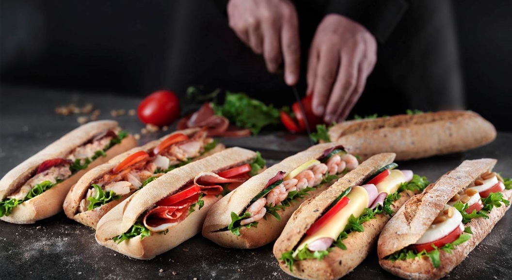 Assorted fresh sandwiches on a dark table with a person slicing tomatoes in the background
