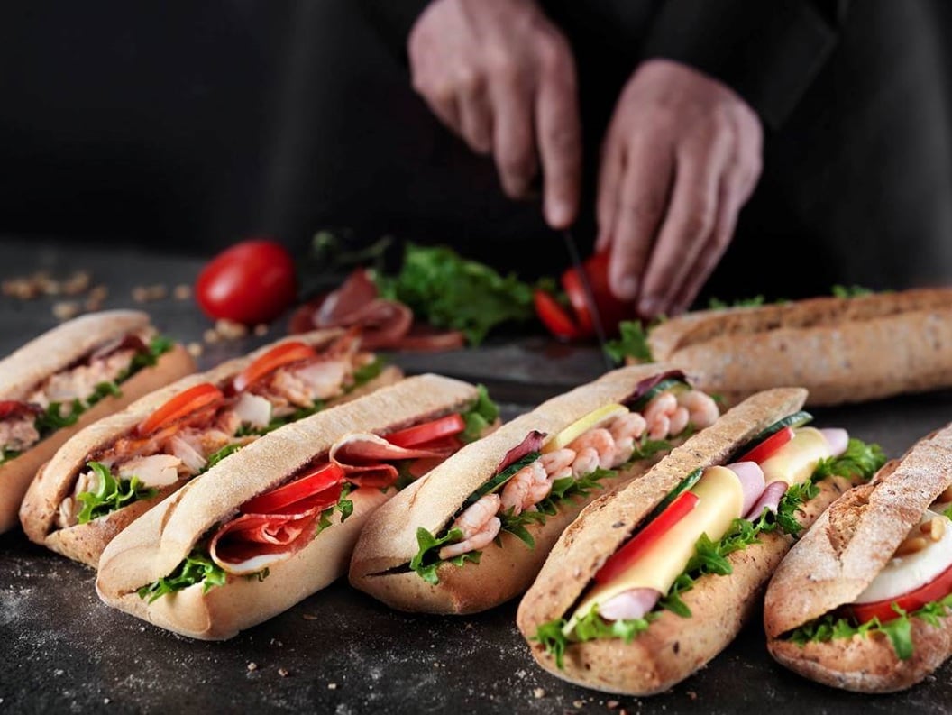 Assorterte ferske smørbrød på et mørkt bord med en person som skjærer tomater i bakgrunnen