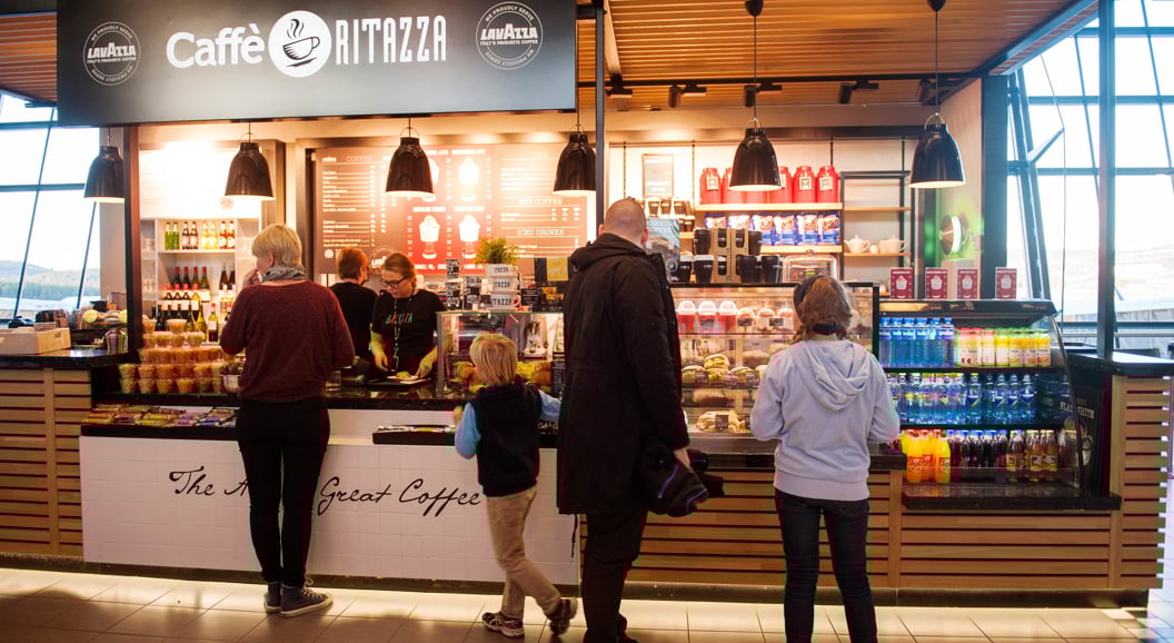 Customers waiting at Caffè Ritazza coffee shop counter, featuring a diverse menu and Lavazza branding, located in an airport terminal