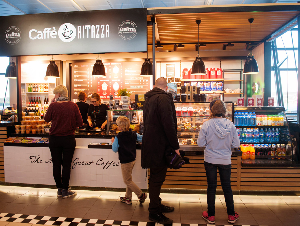 Customers waiting at Caffè Ritazza coffee shop counter, featuring a diverse menu and Lavazza branding, located in an airport terminal