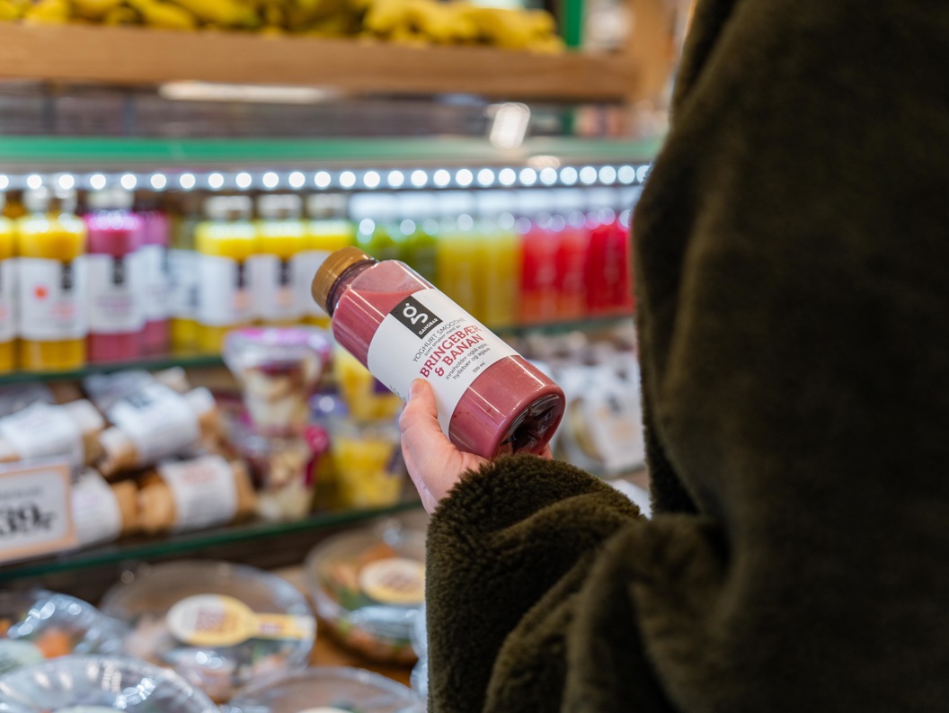 Person som holder en flaske rosa smoothie i en dagligvarebutikk foran en utstilling av ulike fargerike flaskedrikker