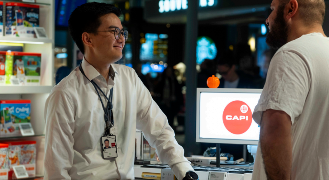 Butikkmedarbeidar møter ein kunde ved skranken i ein elektronikkbutikk, med produktutstilling og ein dataskjerm med Capi-logo i bakgrunnen