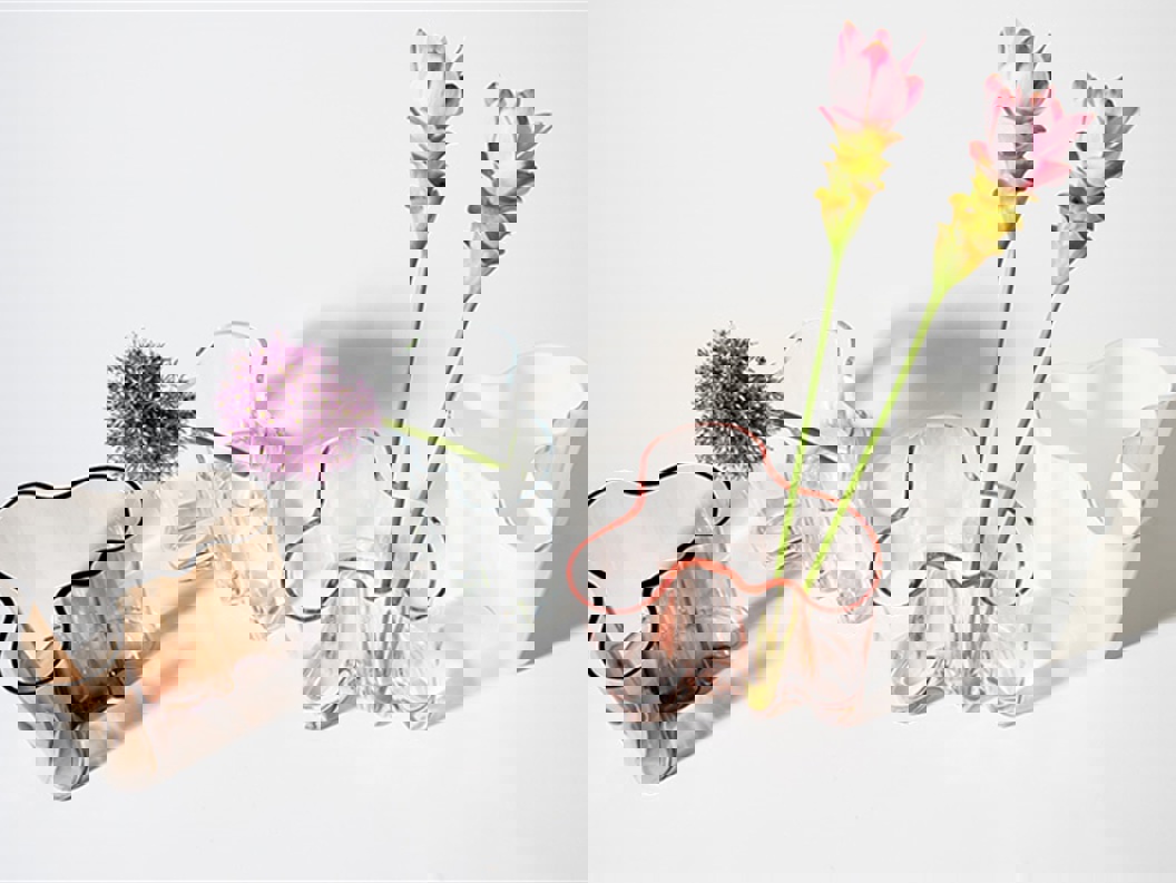 Collection of uniquely shaped glass vases with varying colors, holding pink and purple flowers, displayed on a white background