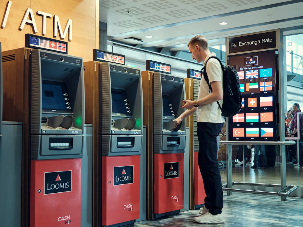 Ein person bruker ein minibank med fleire valutaalternativ ved sidan av ein skjerm som viser valutakursar i ein moderne flyplasshall