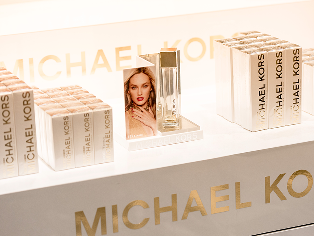 Visning av Michael Kors "Sexy Amber" parfyme med merkede emballasjebokser ryddig arrangert på en butikkhylle