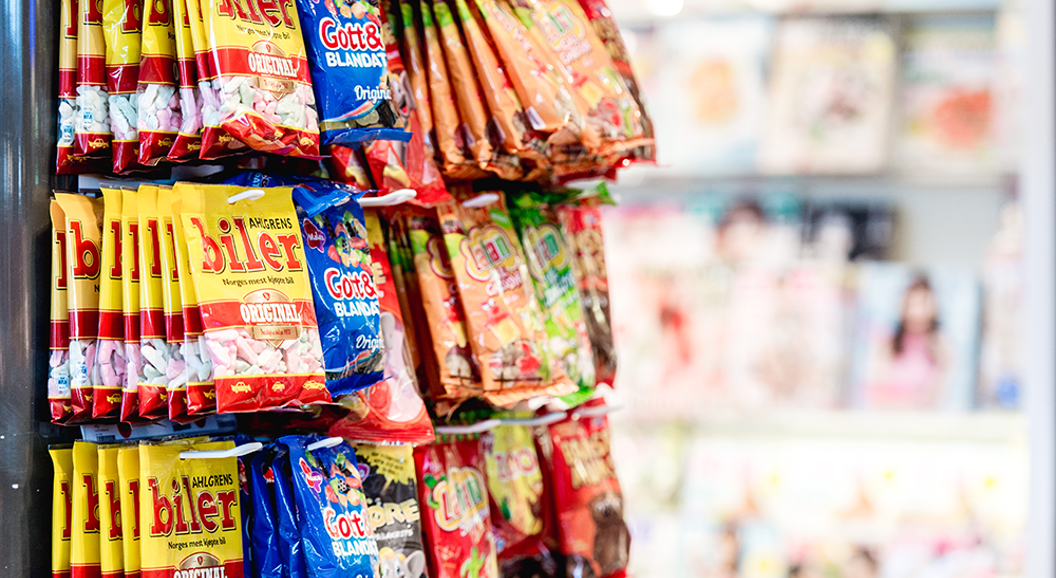 Candy display rack featuring various colorful packaged snacks in a store aisle, with blurred magazine covers visible in the background