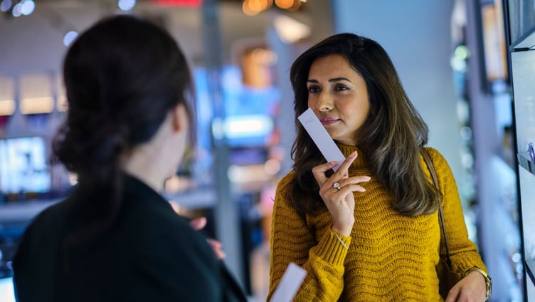 Kvinne i gul genser tester parfyme i en butikk, holder en duftstripe og snakker med en ansatt