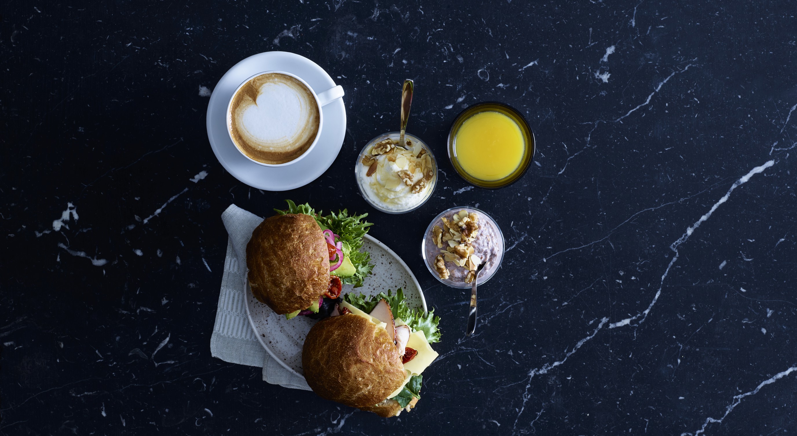 Frokost anrettet på et mørkt marmorbord med en cappuccino, fersk appelsinjuice, en tallerken med smørbrød, og to kremete yoghurtparfaiter