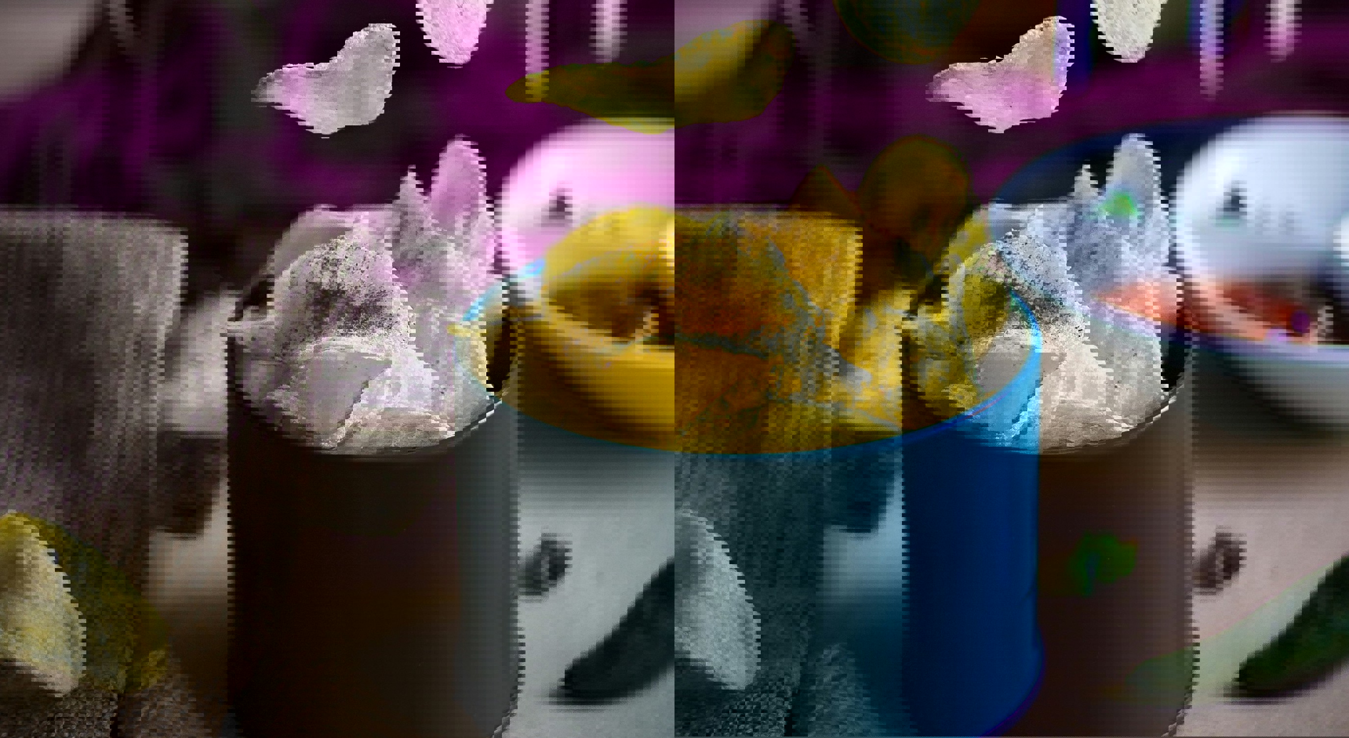 Potetgull i en blå bolle med chips flytende i luften, omgitt av grønne paprikaer og dip i bakgrunnen