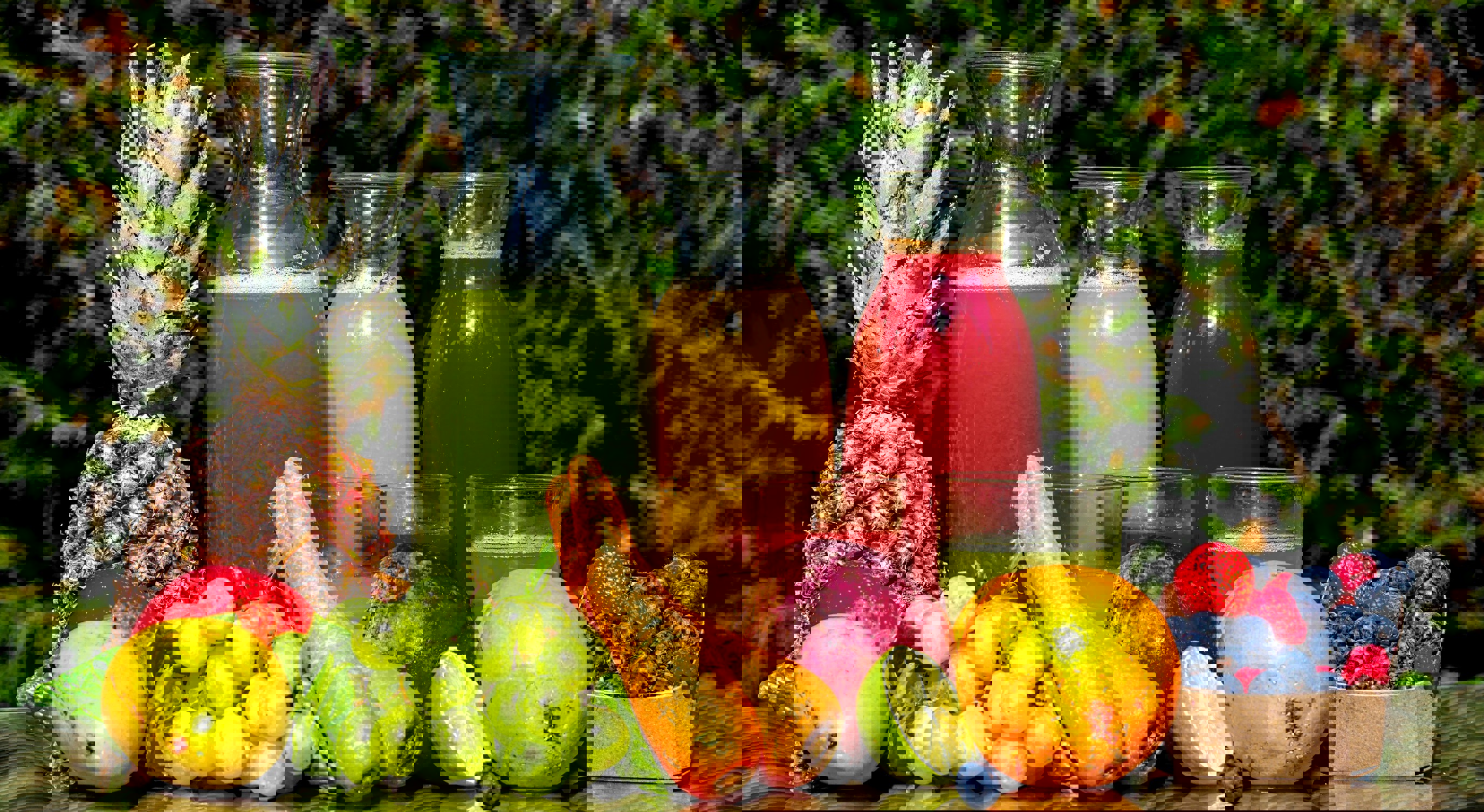 Assorterte ferske frukt- og grønnsaksjuice i glassmugger omgitt av druer, gulrøtter, ananas, lime og bær på en reflekterende overflate med en grønn bladbakgrunn