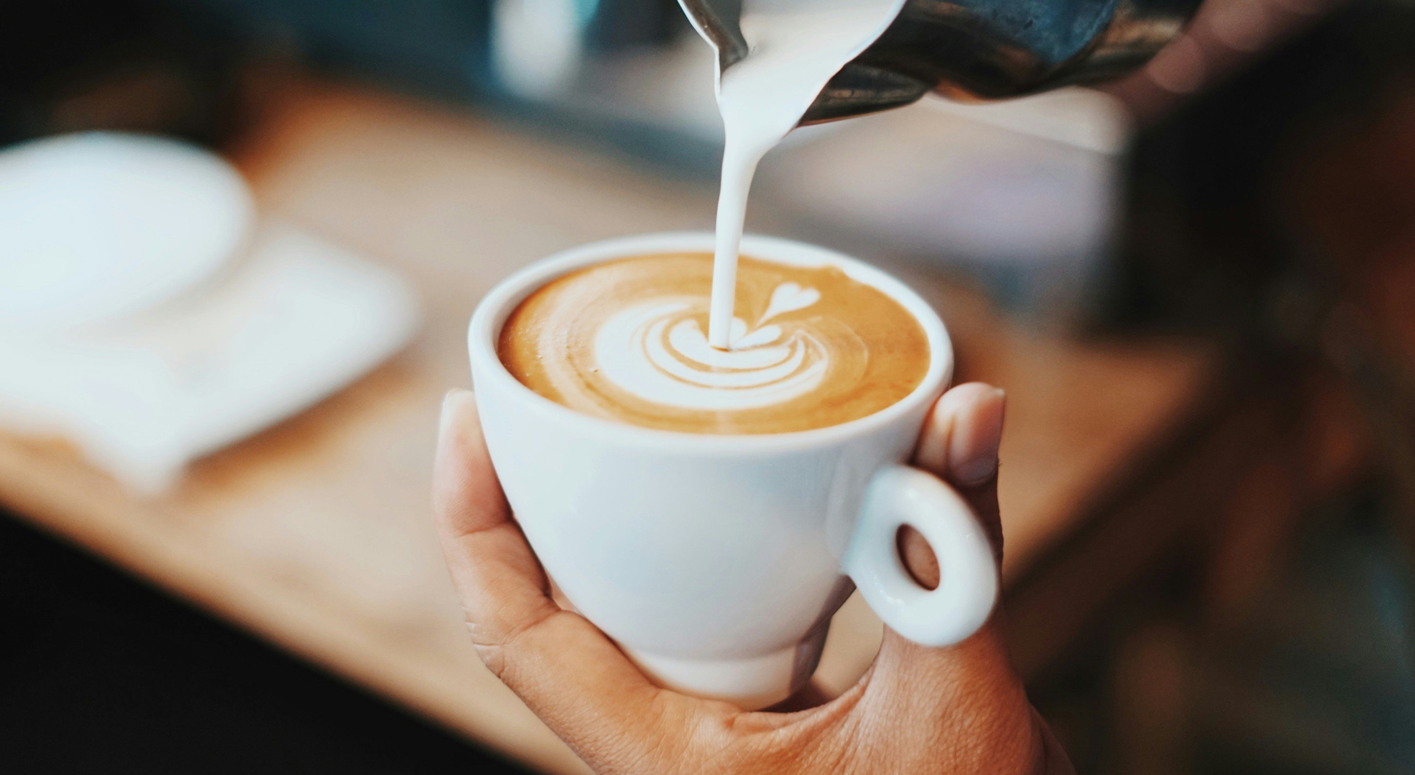 Barista pouring latte art into a cup of coffee, creating a heart shape design, held in a person's hand