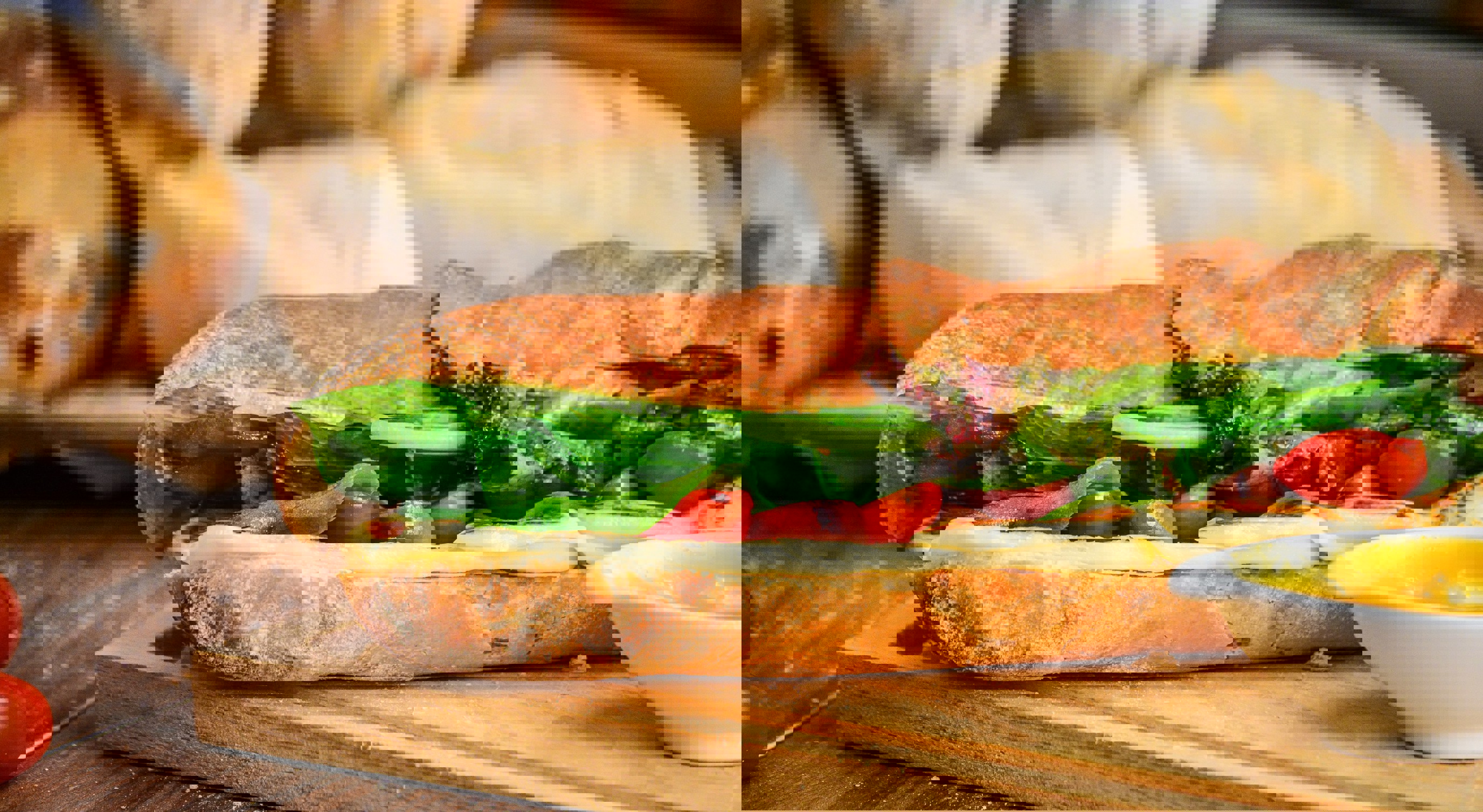 Fresh sandwich with lettuce, tomato, cheese on a crusty baguette, served on a wooden board with a side of mustard, with bread loaves in the background