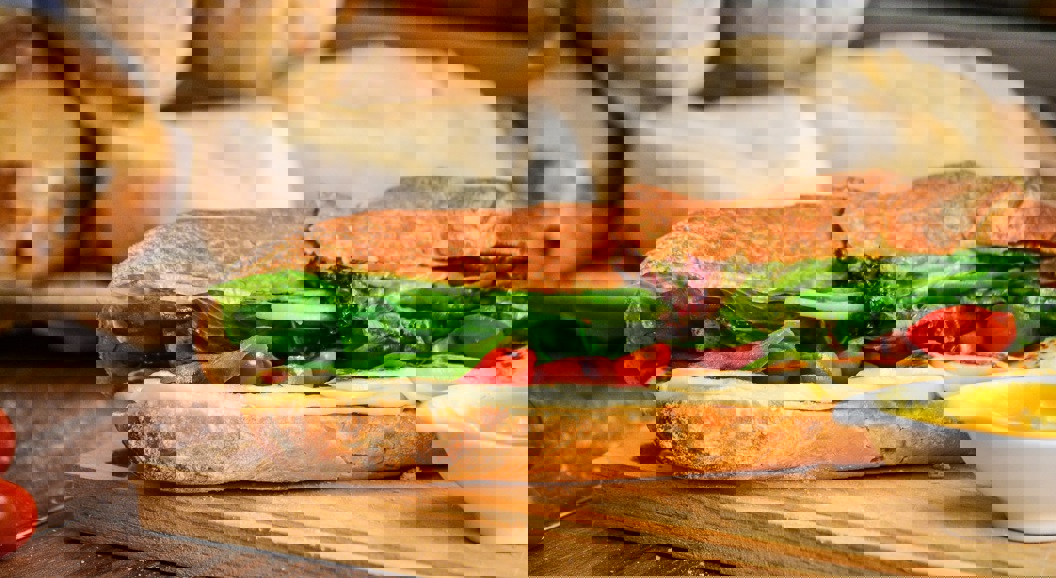 Fersk sandwich med salat, tomat, ost på en sprø baguette, servert på et trebrett med en side av sennep, med brød i bakgrunnen