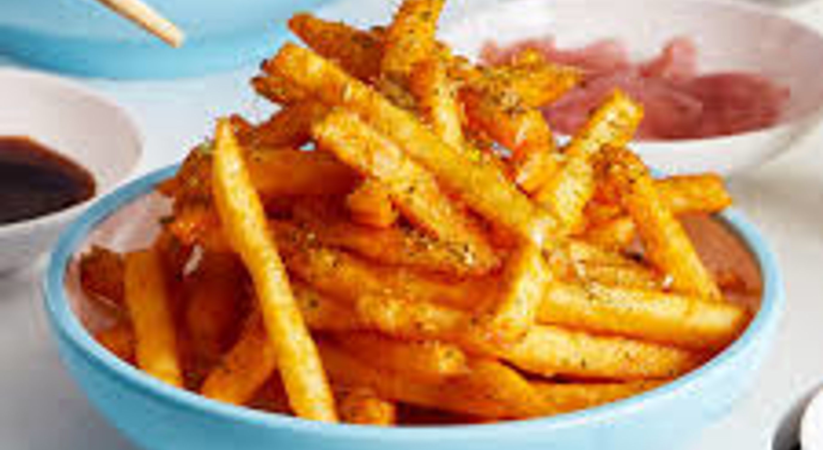 Skål med sprø pommes frites krydret med urter, omgitt av asiatiske retter og tilbehør på et bord