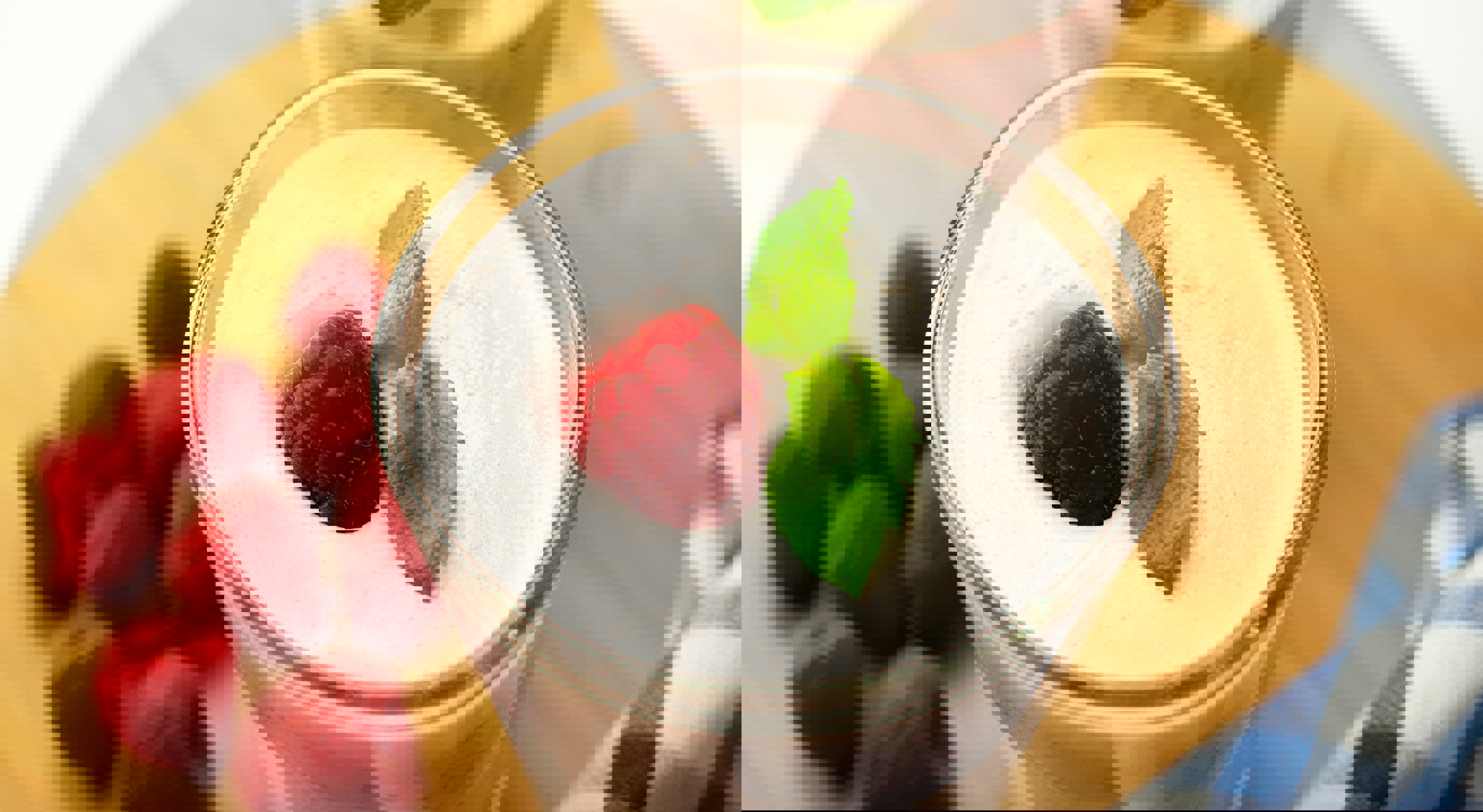 Kremet bringebærmousse i et klart glass toppet med et friskt bringebær og mynteblader på en treoverflate, omgitt av spredte bringebær og en blå rutete klut