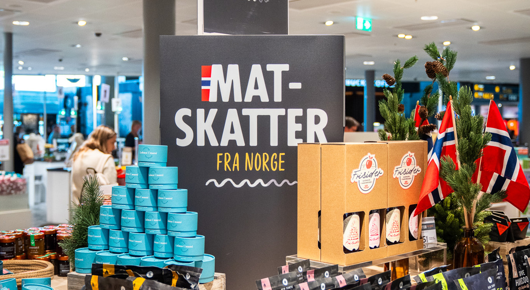 Norsk matutstilling i en butikk som viser et utvalg av pakkede varer, inkludert müsli og syltetøy, med skilt merket "Mat-Skatter fra Norge" og små norske flagg til dekorasjon