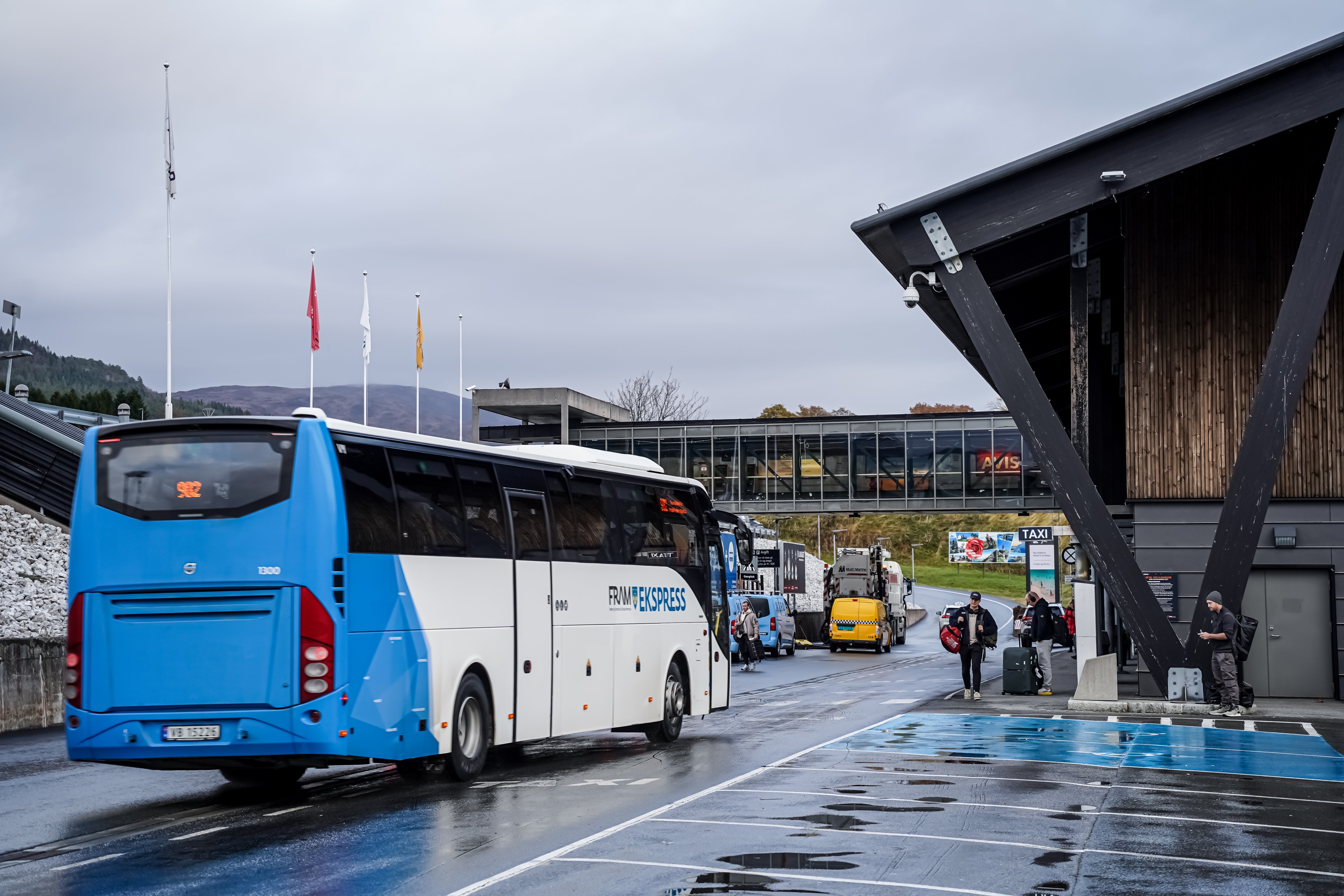 Blå buss utanfor ein flyplassterminal i Noreg, med passasjerar som stig av og nokre står med bagasje. Moderne bygning og parkeringsplass i bakgrunnen. Grå himmel og fuktig ver.