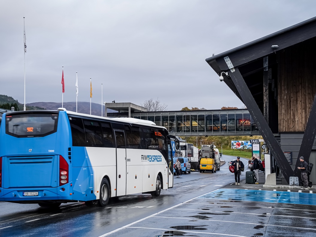 Buss ved terminal med reisende i høstvær, skilt mot taxiholdeplass i bakgrunnen.