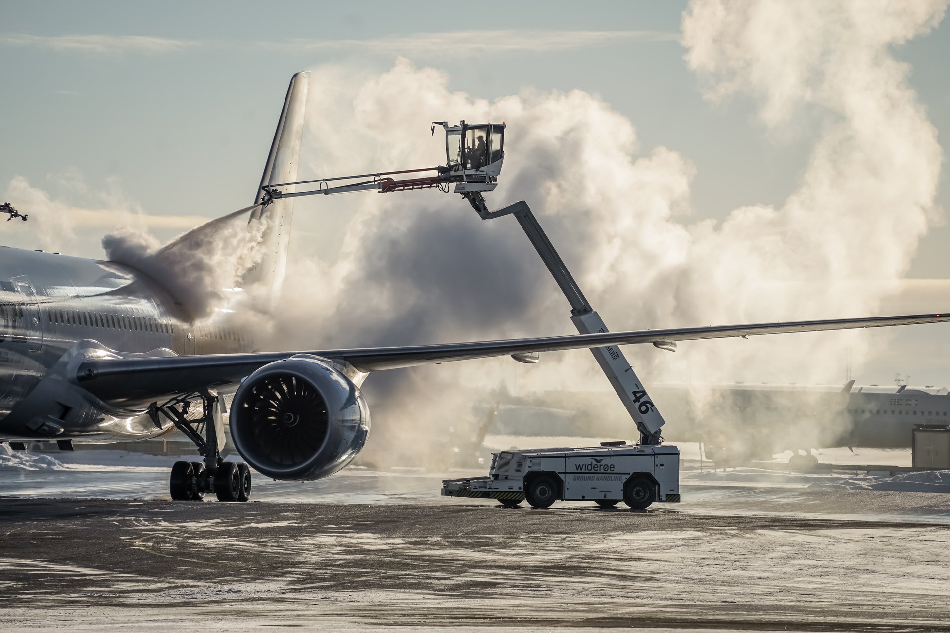 Et passasjerfly blir av-iset på en flyplass, med damp som stiger opp i kulden. En de-isebil er i aksjon under klar himmel.