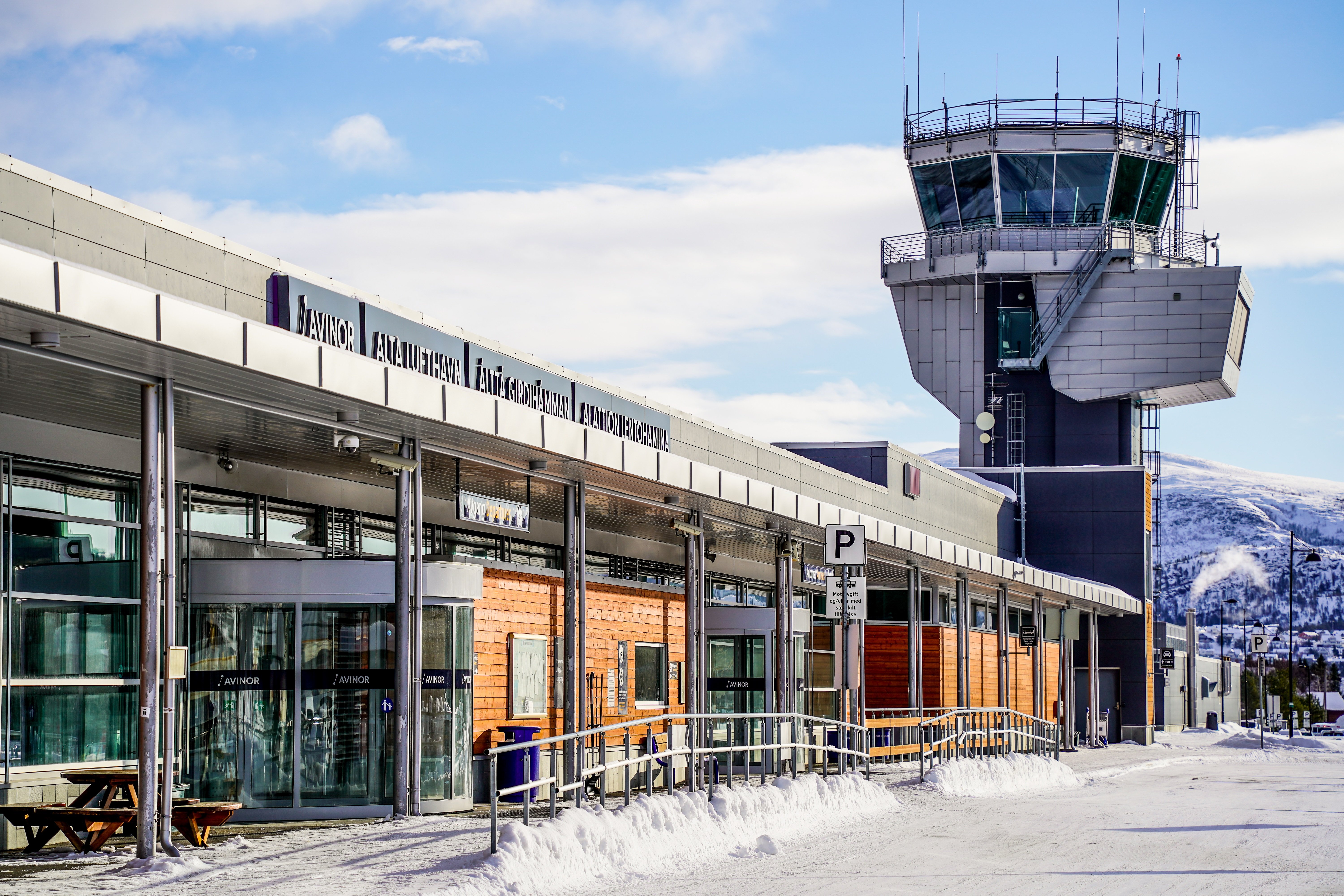 Flyplass med kontrolltårn i vinterlandskap, med skilt for Avinor Alta lufthamn, snødekt bakke og klar blå himmel.