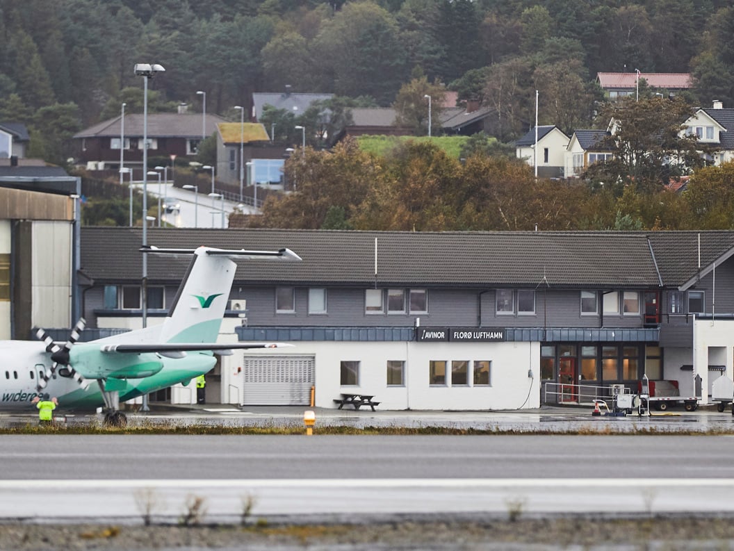 Widerøe-fly ved Florø lufthavn foran terminalbygningen, omgitt av skog og boliger i bakgrunnen.