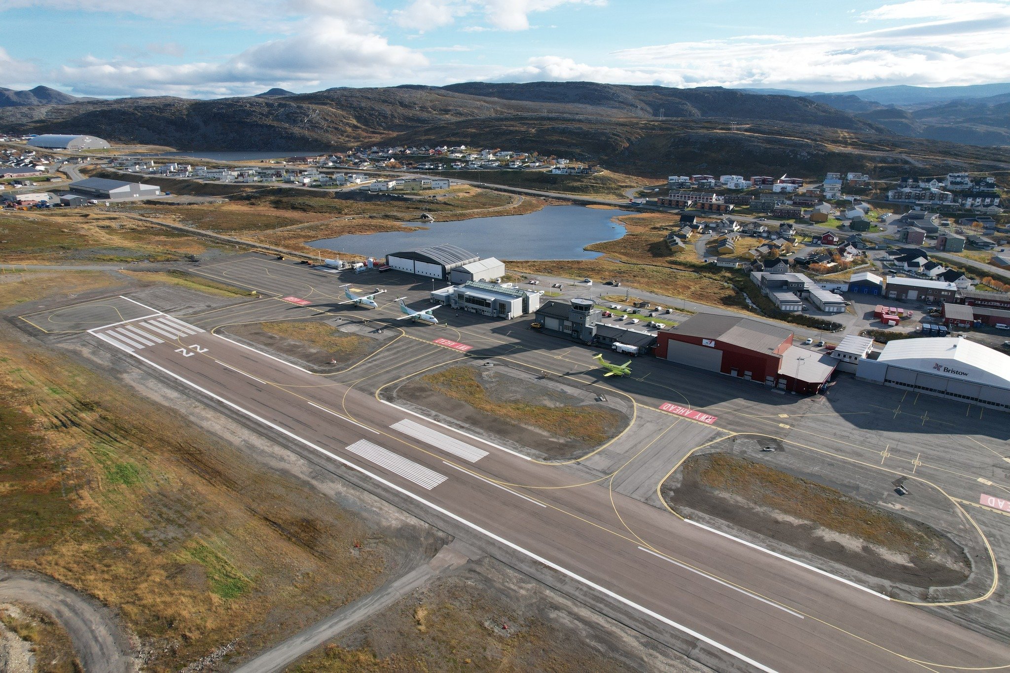 Aerial photo of Hammerfest Airport with runway and terminal building surrounded by hilly terrain and a village in the background.