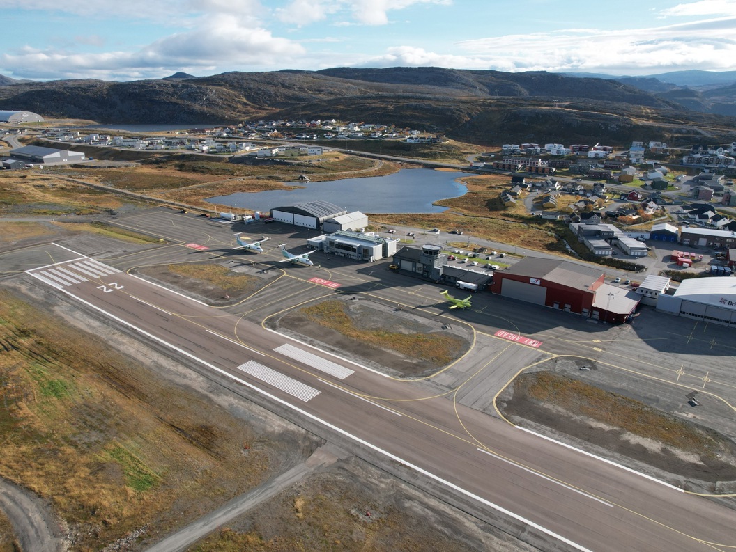 Flyfoto av Hammerfest flyplass med rullebane og terminalbygg omgitt av kupert landskap og en bygd i bakgrunnen.