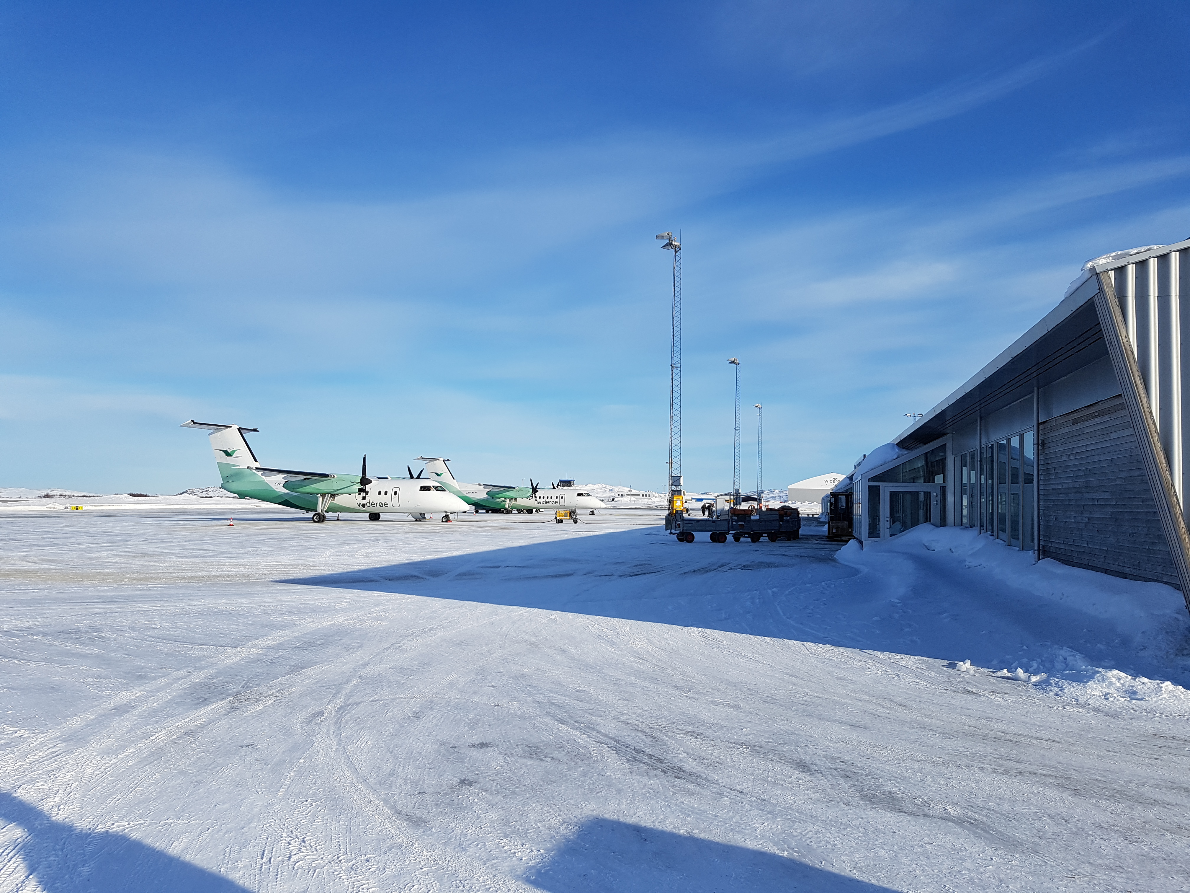 To fly på ein snødekt rullebane ved Kirkenes flyplass i Noreg under klar blå himmel. Bygningar i nærleiken viser moderne arkitektur.