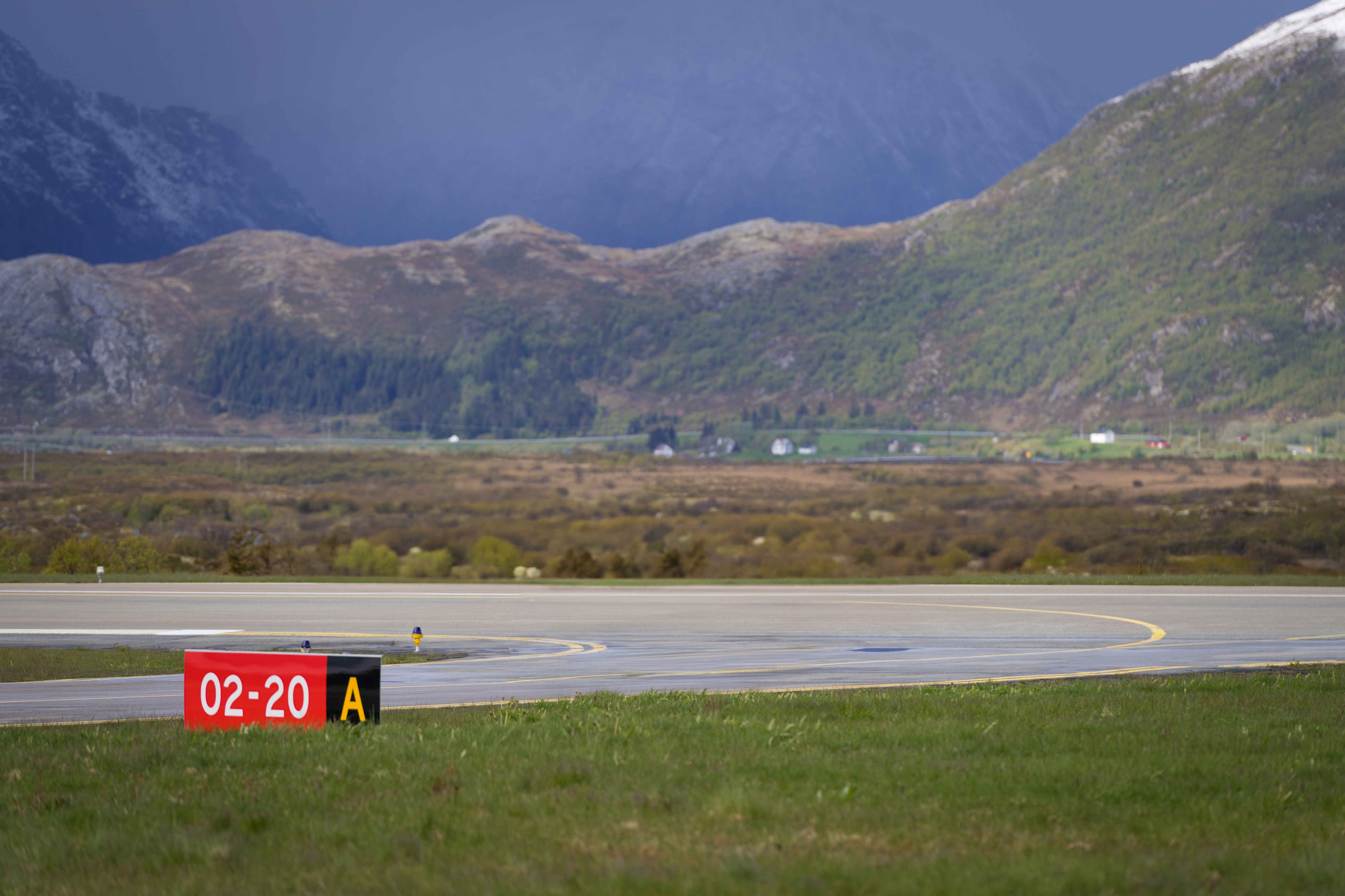 Flyplassrullebane med skilt 02-20 A, omgjeven av norsk fjellandskap og grøne dalar under overskya himmel.