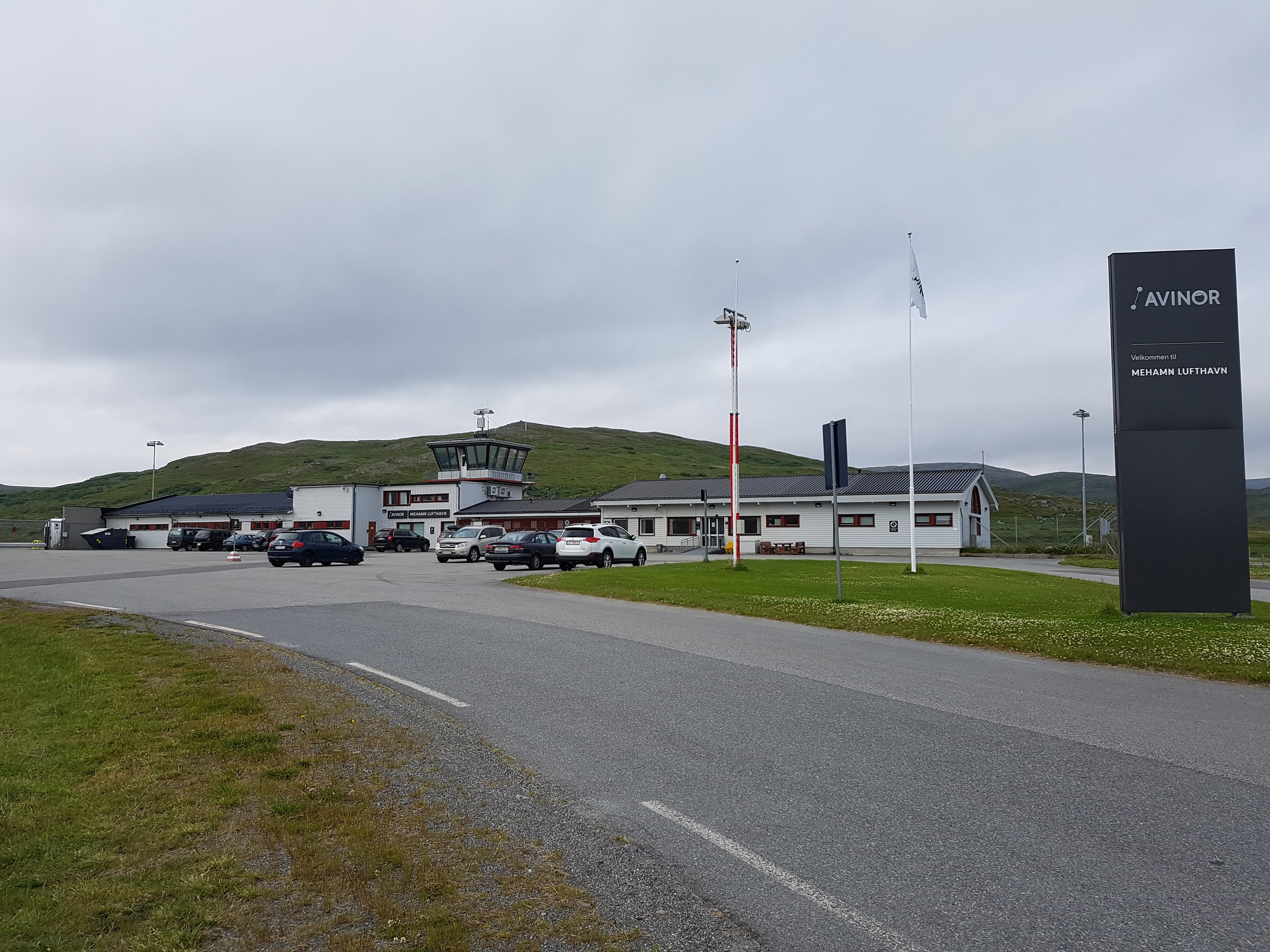 Mehamn Airport in Norway, with parked cars and a control tower, surrounded by green hills under a cloudy sky.
