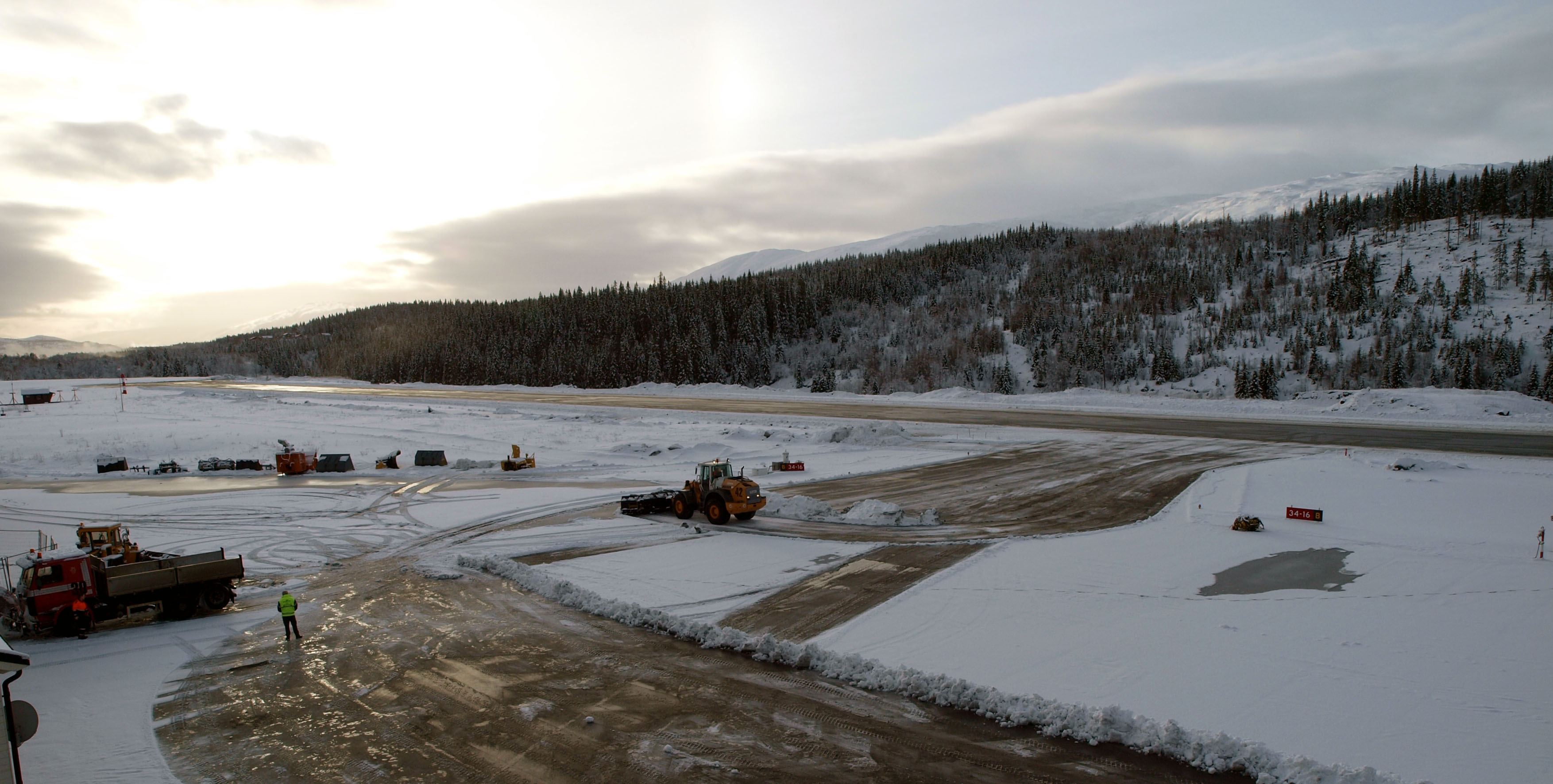 Snødekt flyplass med rullebane i vintersol, omgitt av skogkledde åsar. Snøryddingsutstyr er i arbeid på plassen.