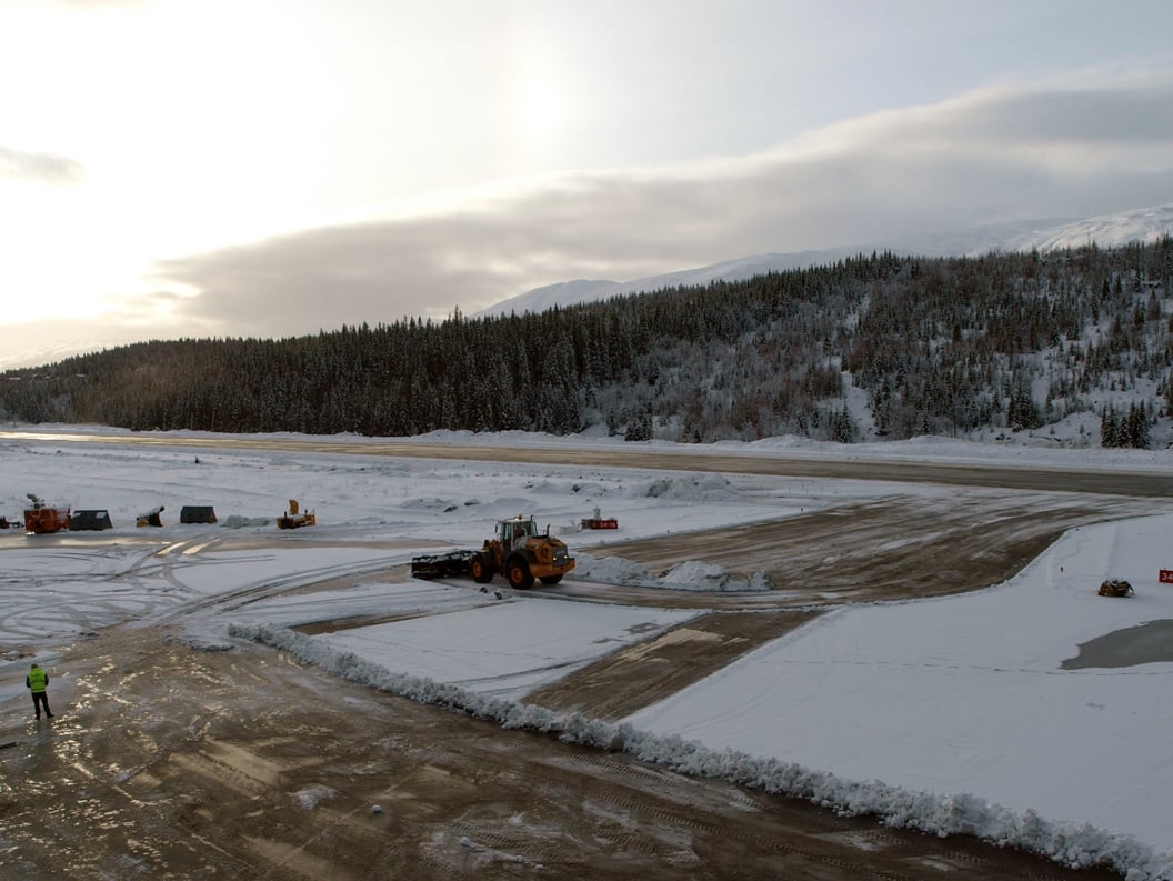 Snødekt flyplass med flystripe i vintersol, omgitt av skogkledde åser. Snøryddingsutstyr er i arbeid på plassen.