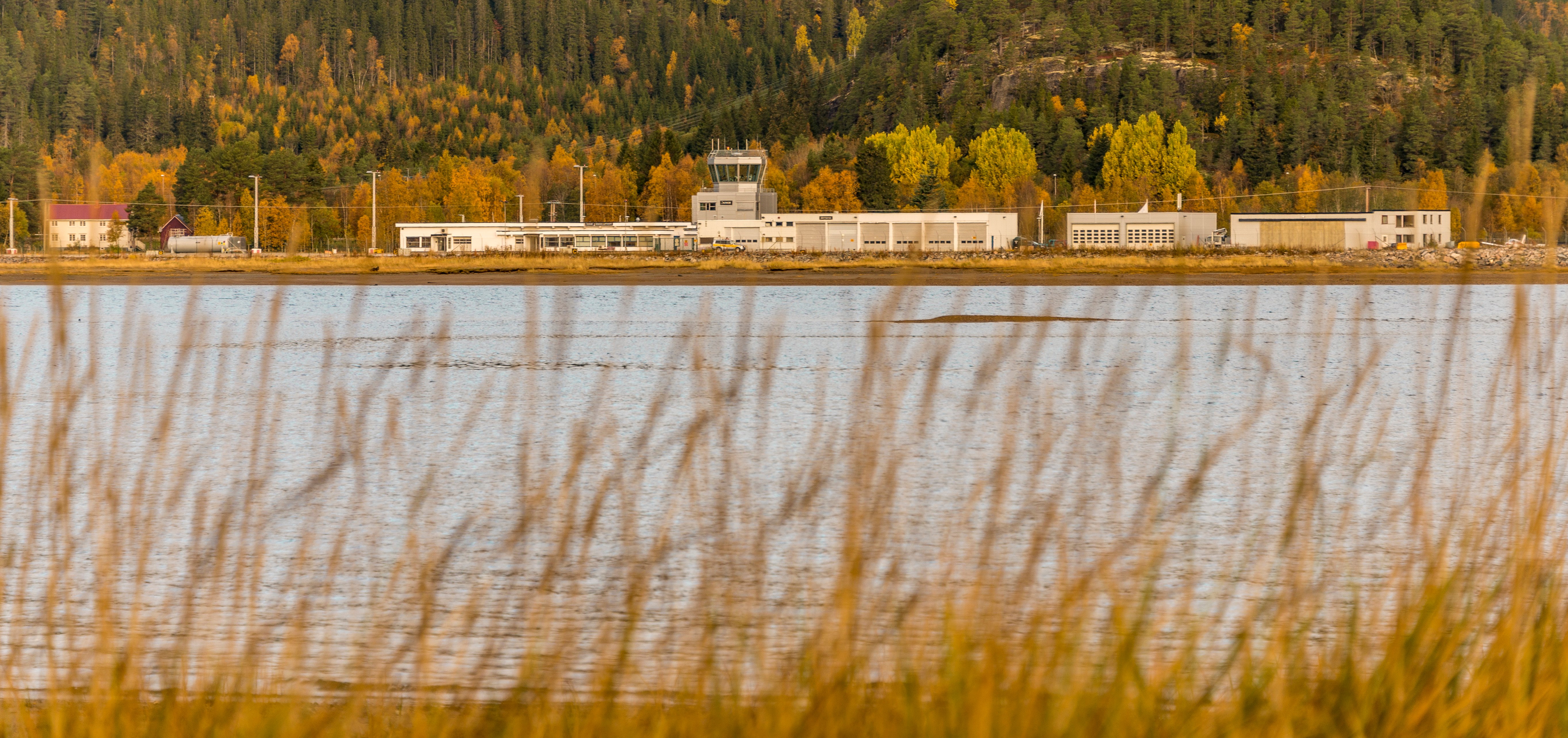 Flyplassbygning ved innsjø om hausten, omringa av haustfarga tre og åsar i bakgrunnen, med siv i forgrunnen.