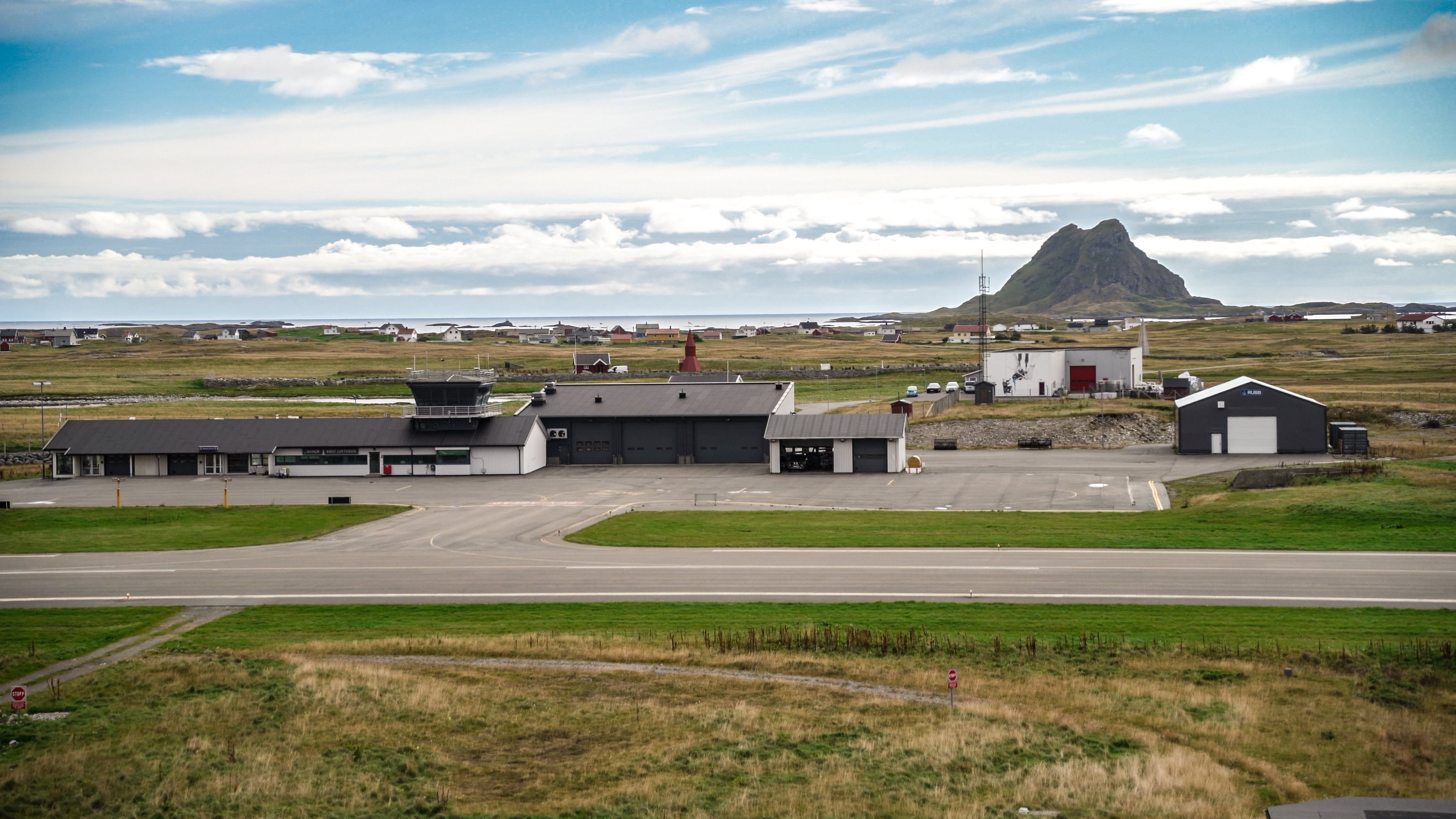 Flyplassbygning med rullebane i eit landleg landskap med fjell i bakgrunnen under ein delvis skya himmel.