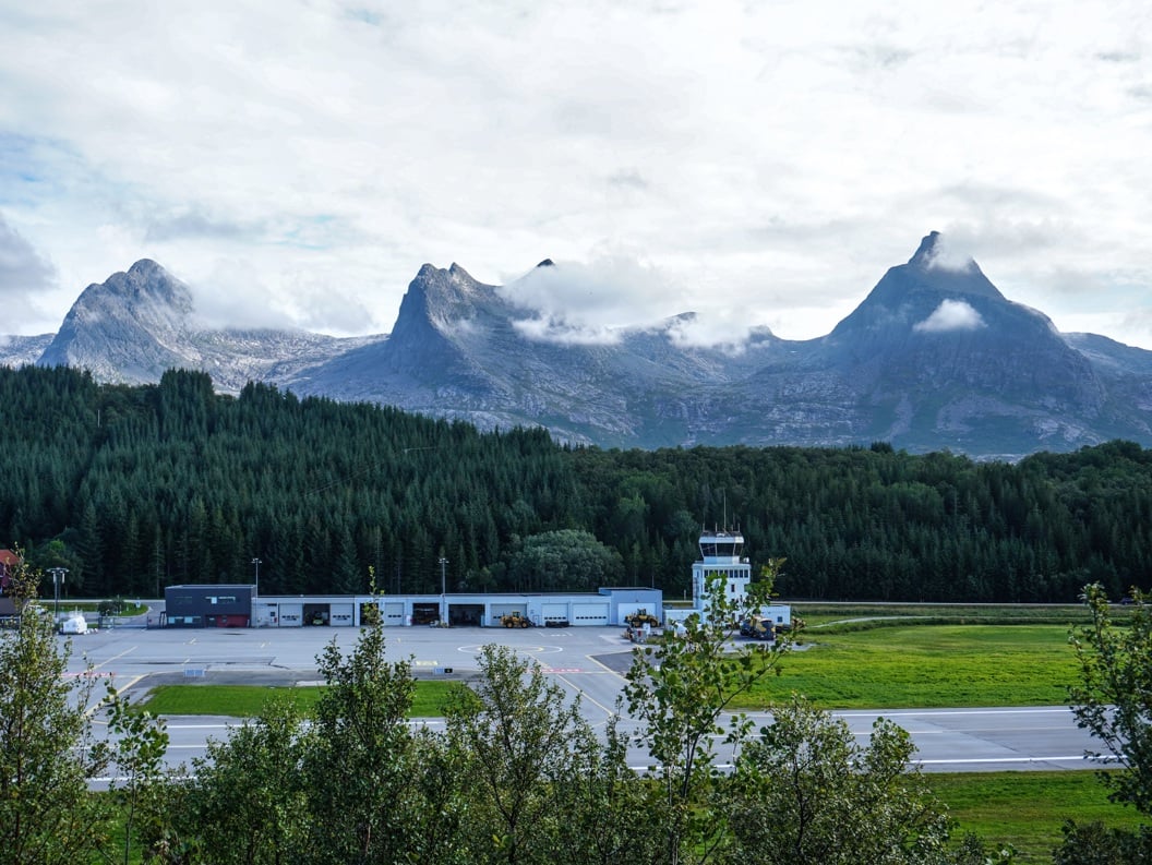 Sandnessjøen flyplass omgitt av skogkledde åser og majestetiske fjell under en overskyet himmel i Norge.