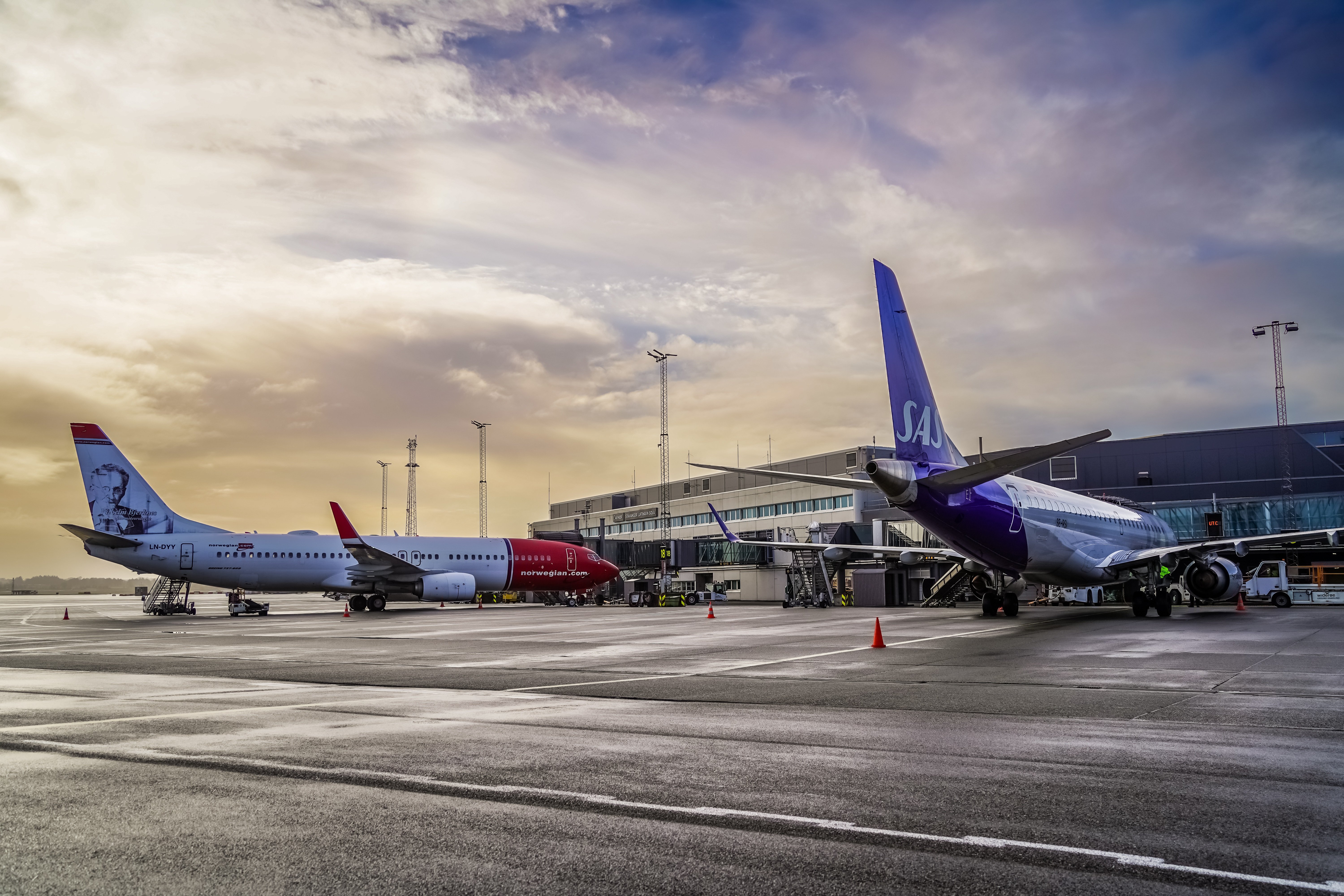 To fly på en flyplass i solnedgangen, et Norwegian-fly og et SAS-fly parkert ved en terminal, med et moderne terminalbygg i bakgrunnen.