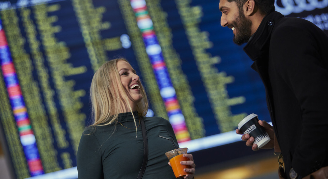 To personar smiler og held drikke i ein flyplass terminal framfor ein avgangstavel