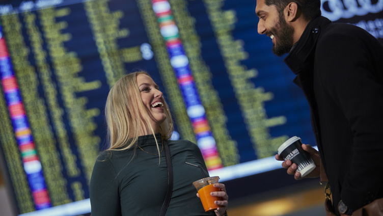 To personar smiler og held drikke i ein flyplass terminal framfor ein avgangstavel