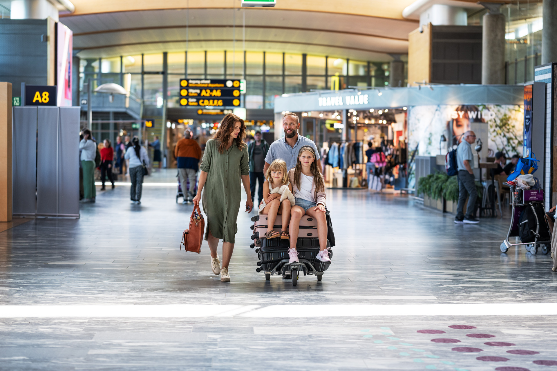 Familie med bagasjevogn på flyplass, blant reisebutikker og avganger.