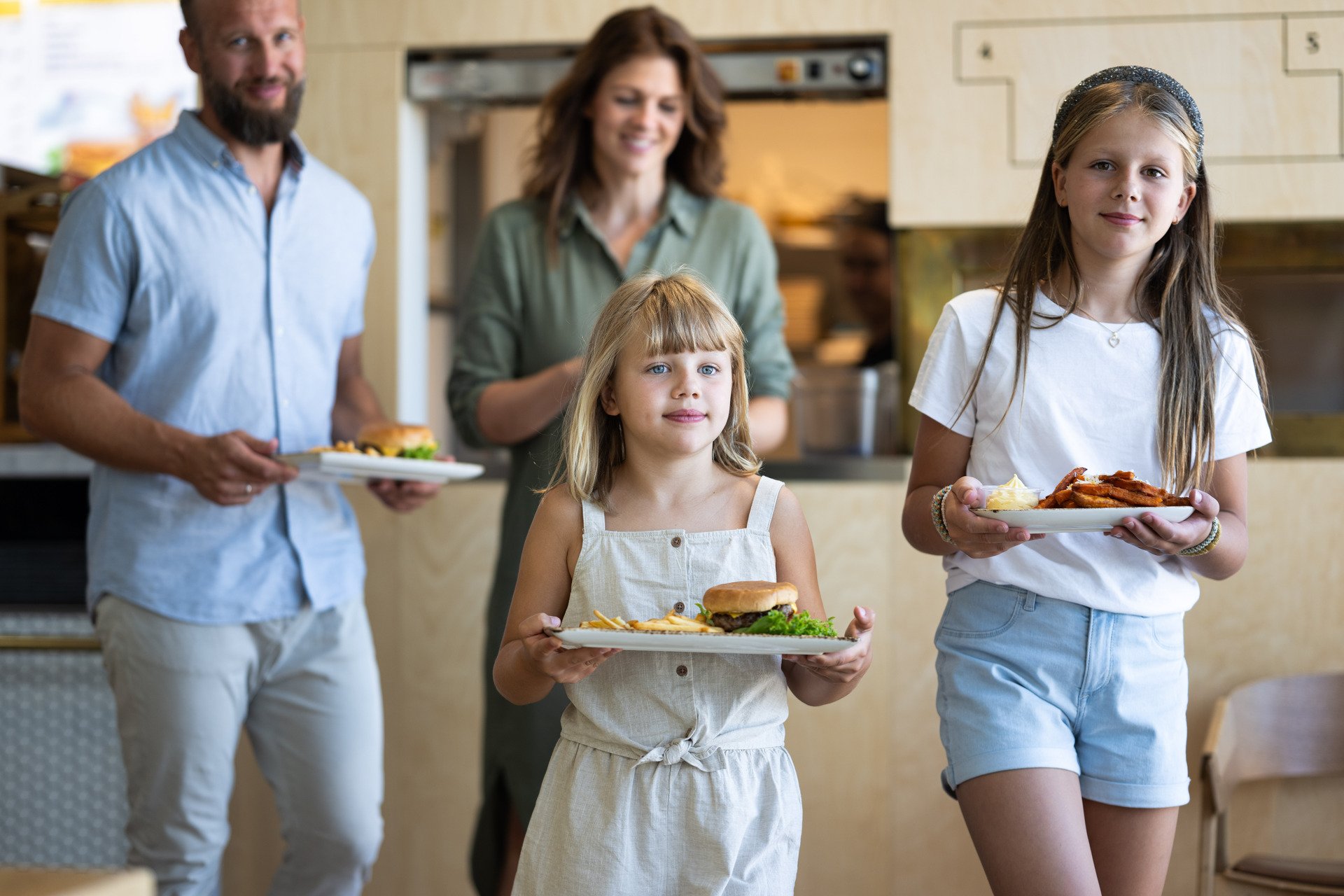 Family with two children enjoying a meal at a restaurant, serving hamburgers and fries. The parents are smiling in the background.