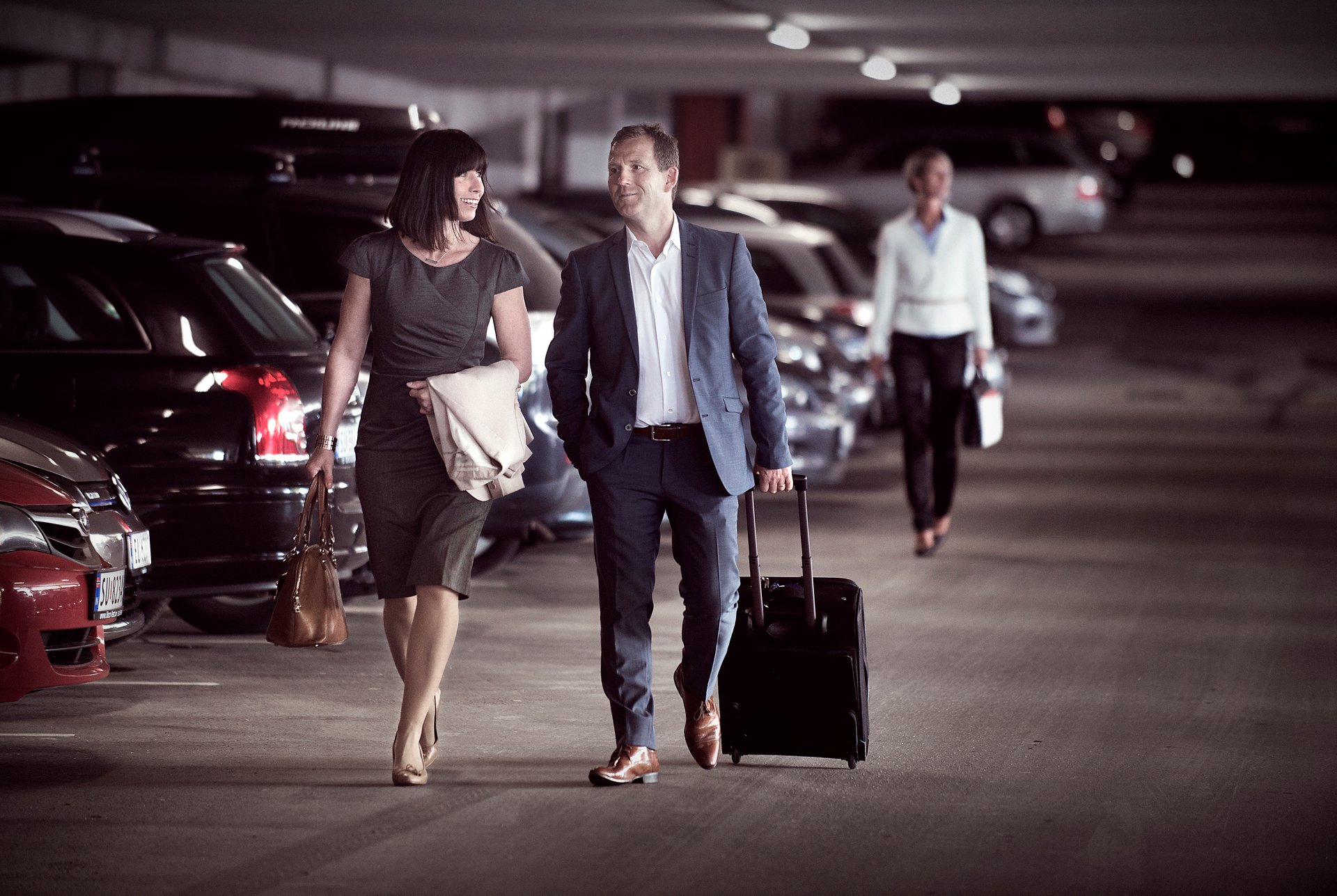 Business people in a parking garage, a man in a suit rolling a suitcase while a woman next to him carries a handbag.