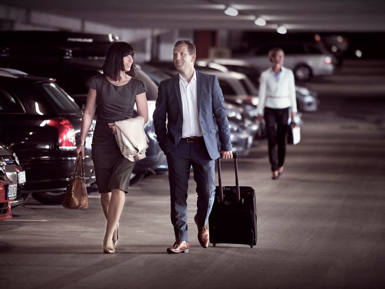 Business people in a parking garage, a man in a suit rolling a suitcase while a woman next to him carries a handbag.