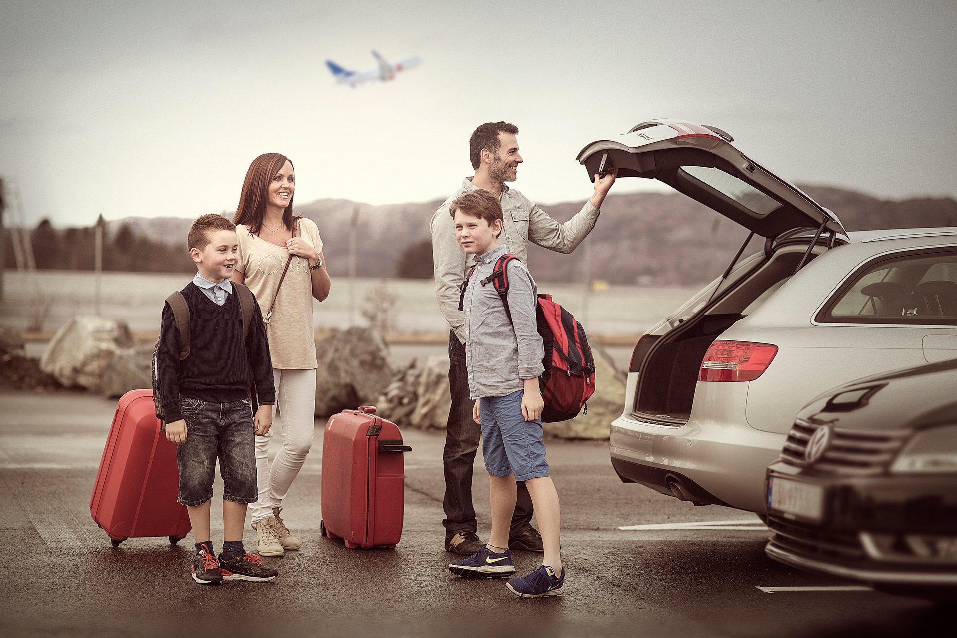 Family of four by car at the airport with suitcases, ready to travel with a plane in the background.
