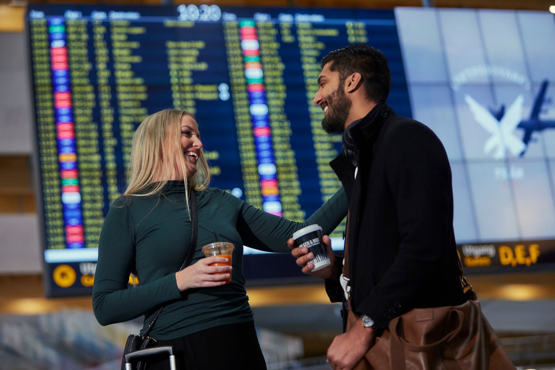 Two people at an airport are smiling and holding drinks, with a departure board visible in the background.