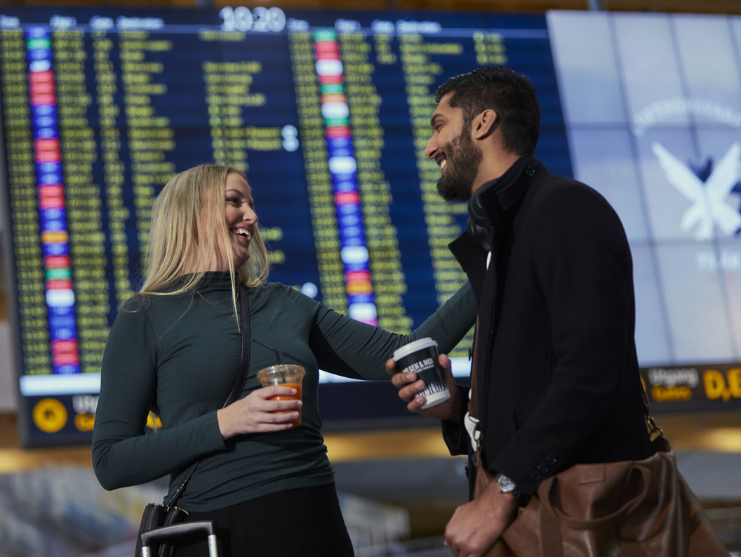 To personar smiler og held drikke framfor ei flyplassevle med flyinformasjon.